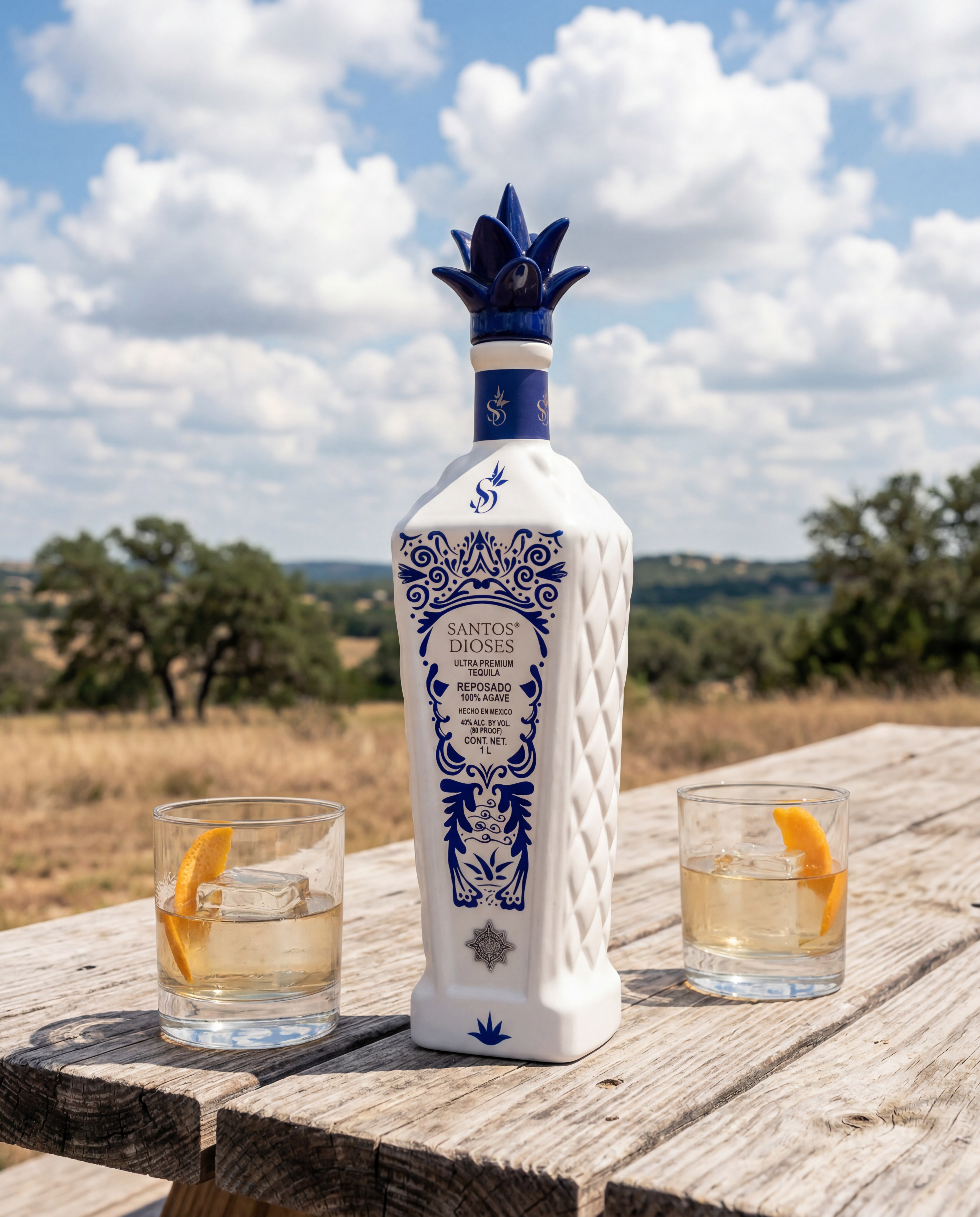 Santos Dioses Reposado bottle hero on a weathered wood plank picnic table in the Texas Hill Country at midday, two rocks glasses flanking the bottle with single clear ice cube and a sliver of orange peel garnish, near-clear pour, rolling oak-studded hills and big cumulus clouds behind, no people